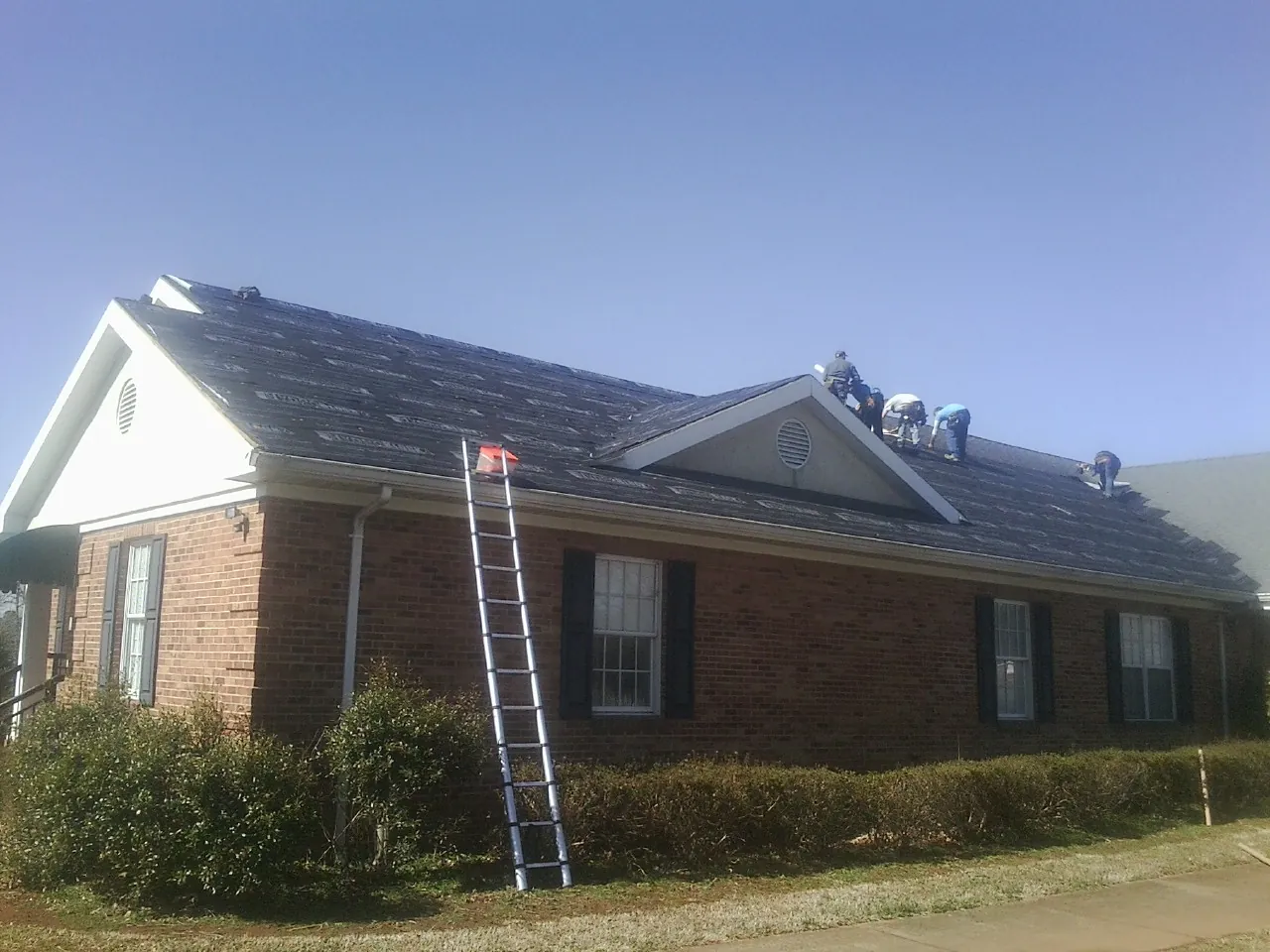 Crew tearing shingles off a brick ranch roof with a ladder set — Emergency Roof Repair work by our crew in Mineville