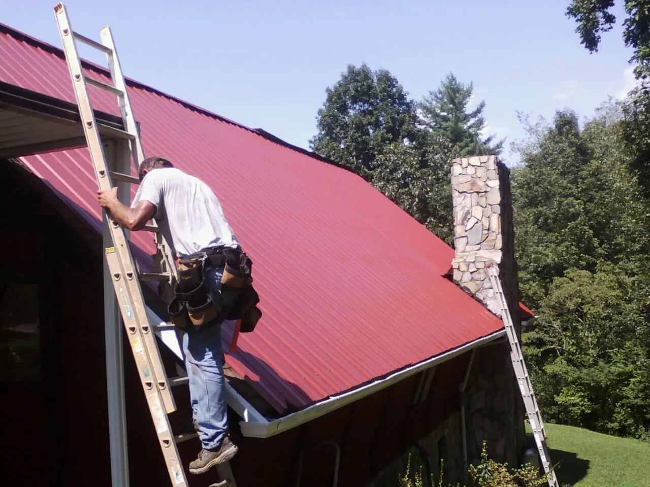 Roofer on a ladder beside a red metal roof and stone chimney — Soffit & Fascia Repair by Roofing Brothers in Mineville